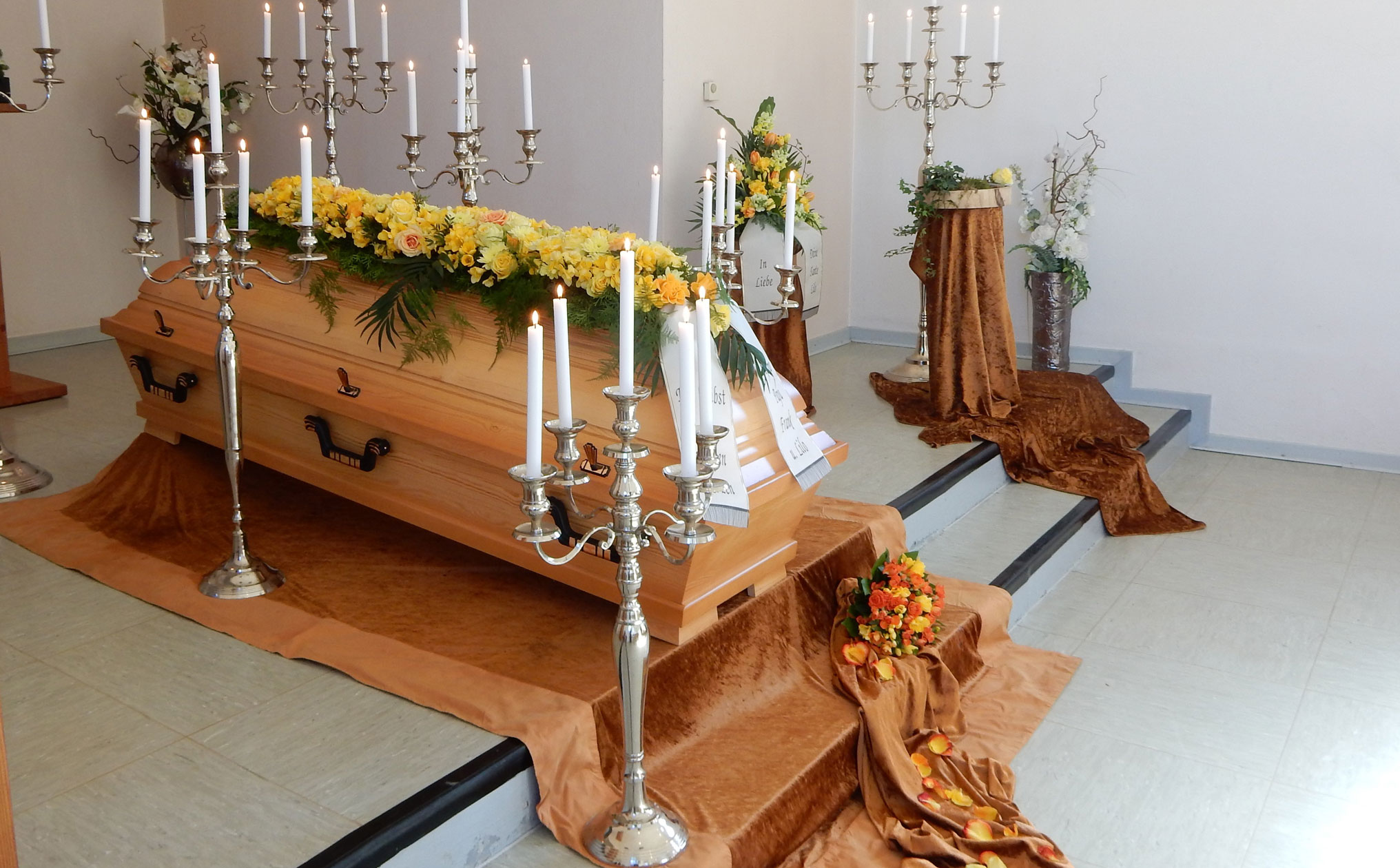 Traditionelle Sargaufbahrung mit gelben Blumen, silbernen Kerzenleuchtern und einer warmen Dekoration für eine feierliche Abschiednahme.