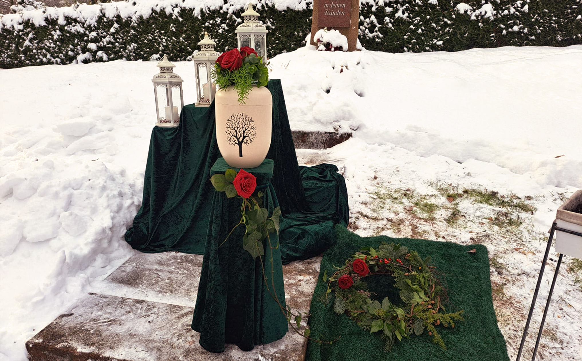 Winterliche Urnenbeisetzung im Schnee, mit einer dekorierten Urne, roten Rosen und Laternen auf einem Friedhof.